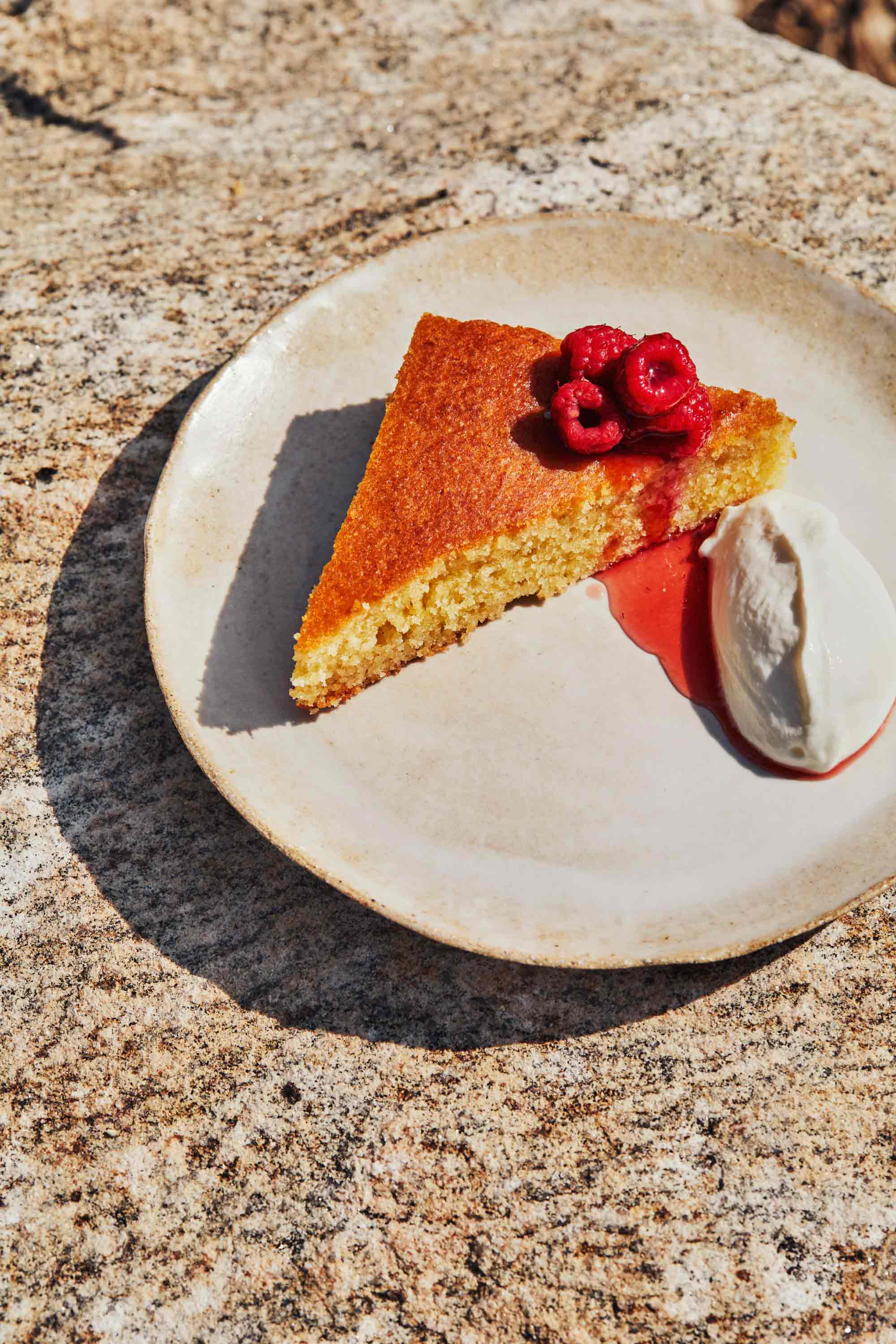 A cake on a beige plate, garnished with a handful of raspberries on top and yogurt whipped cream to the side.