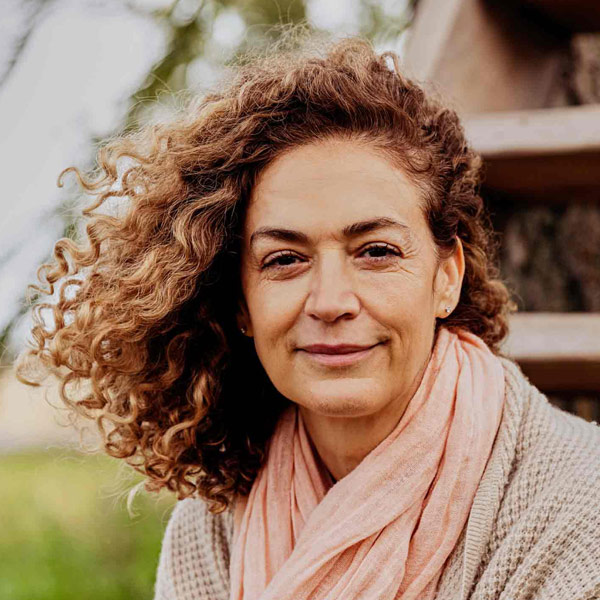 A portrait of a smiling Turkish woman with curly hair blown to one side, wearing a pink scarf and beige cardigan.