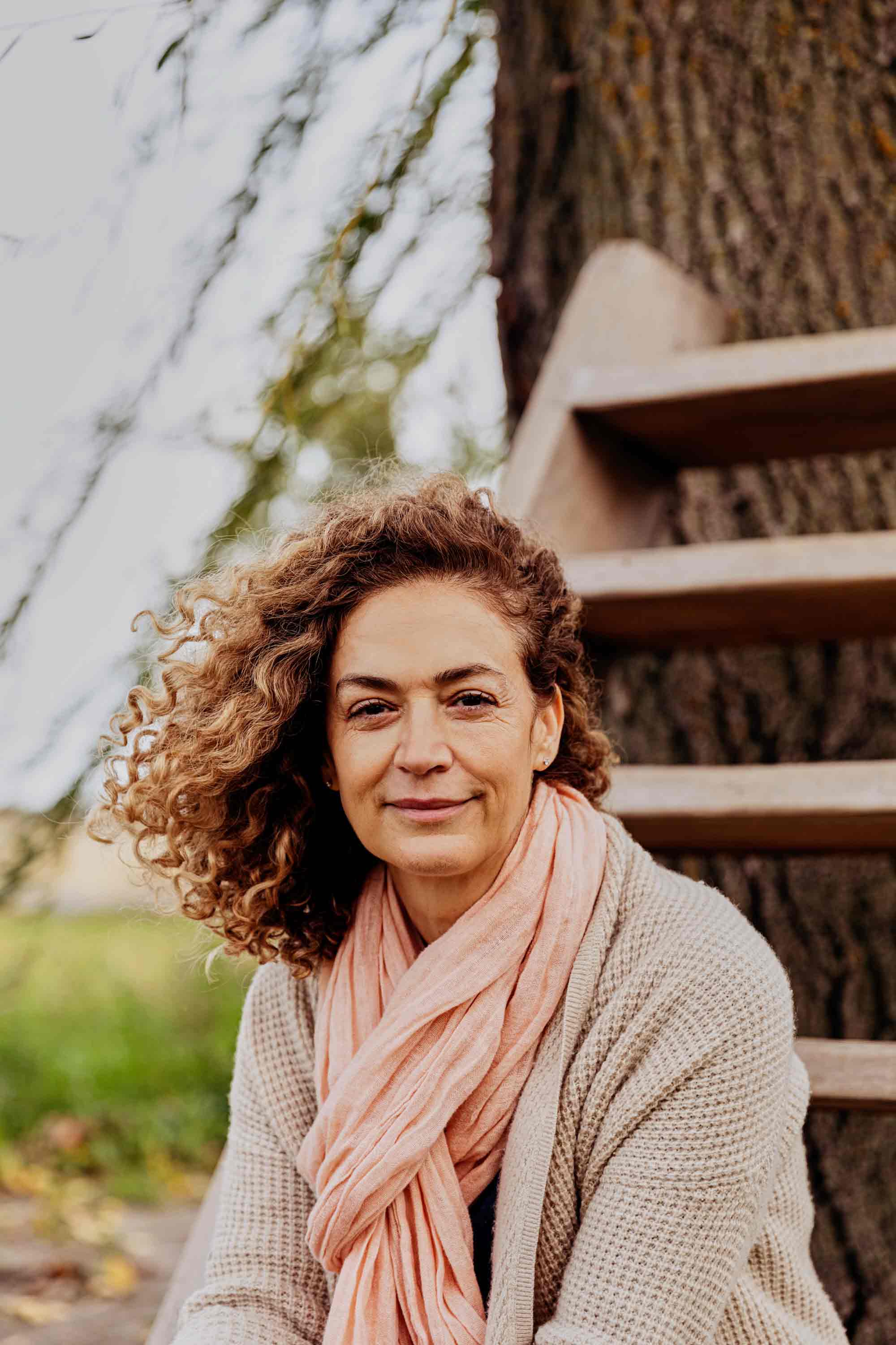 A portrait of a smiling Turkish woman with curly hair blown to one side, wearing a pink scarf and beige cardigan.