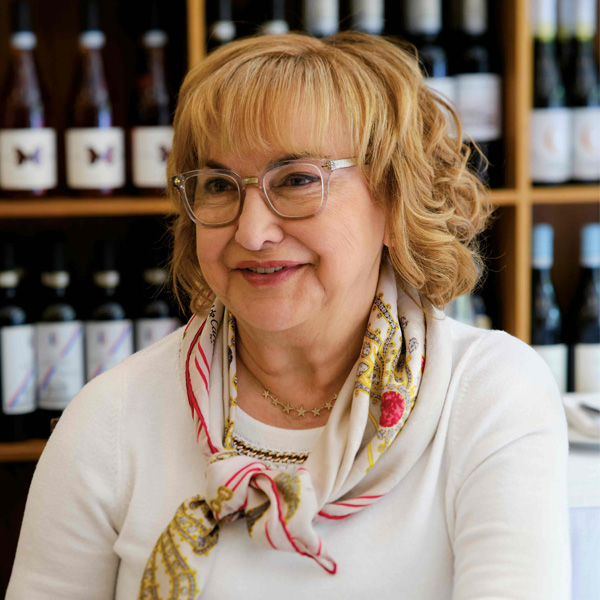 A portrait of a smiling Italian woman wearing a silk scarf and white shirt, with wine bottles in the background.