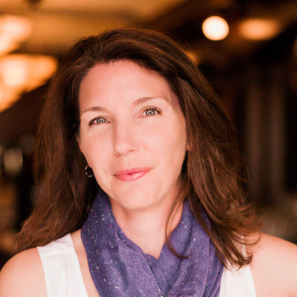 A portrait of a smiling white woman wearing a white tank top and purple scarf, leaning against a bar.