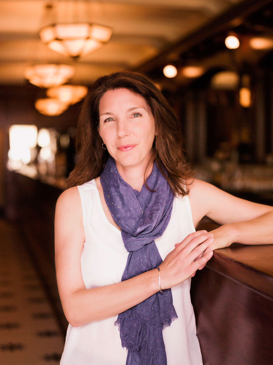 A portrait of a smiling white woman wearing a white tank top and purple scarf, leaning against a bar.