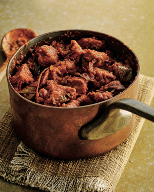 A pot with pork vindaloo in it, resting on a fringed kitchen towel with a serving spoon beside it.