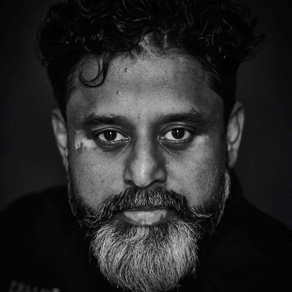 A black and white portrait of an Indian man with short, curly hair and a trim, greying beard looking into the camera.