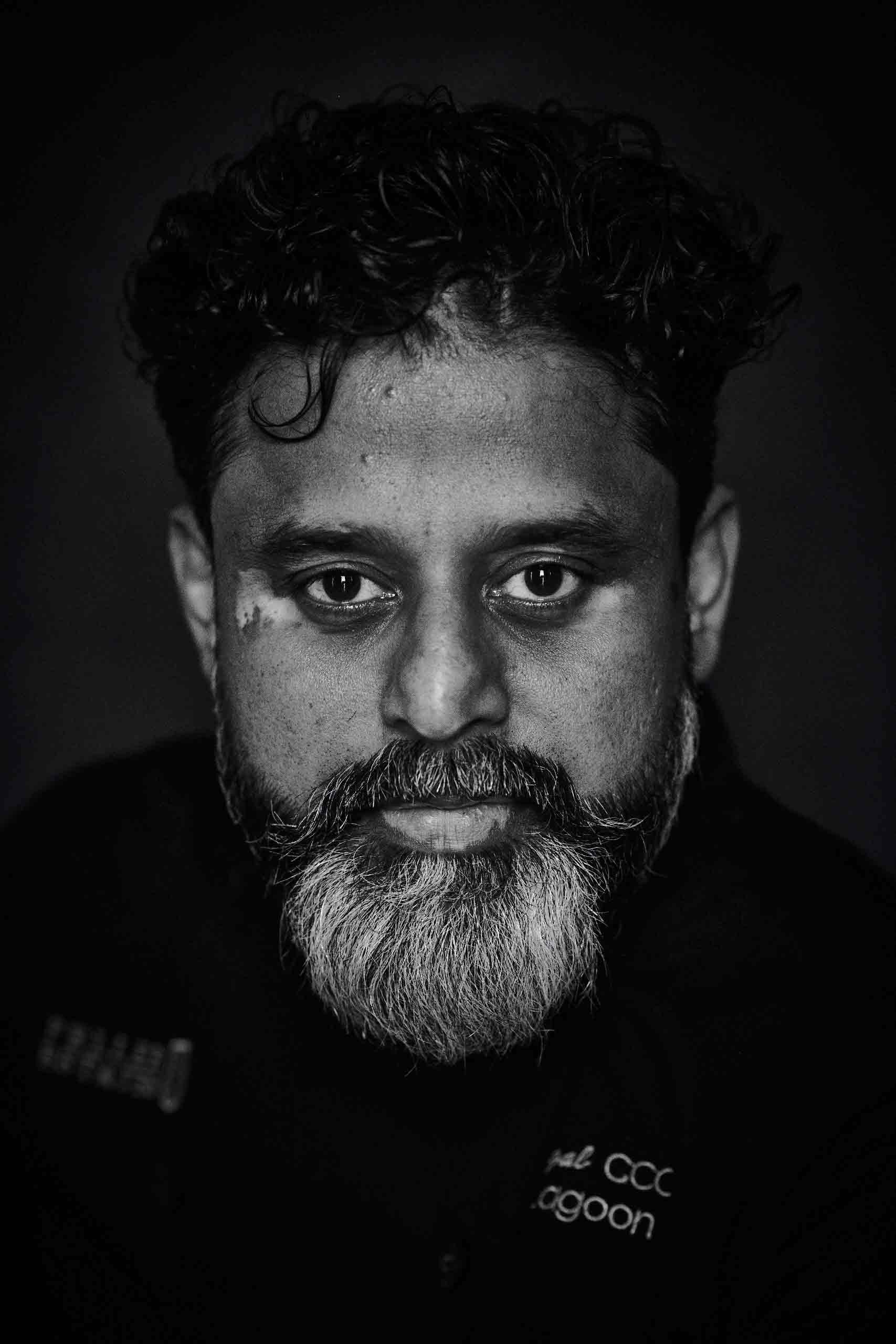 A black and white portrait of an Indian man with short, curly hair and a trim, greying beard looking into the camera.