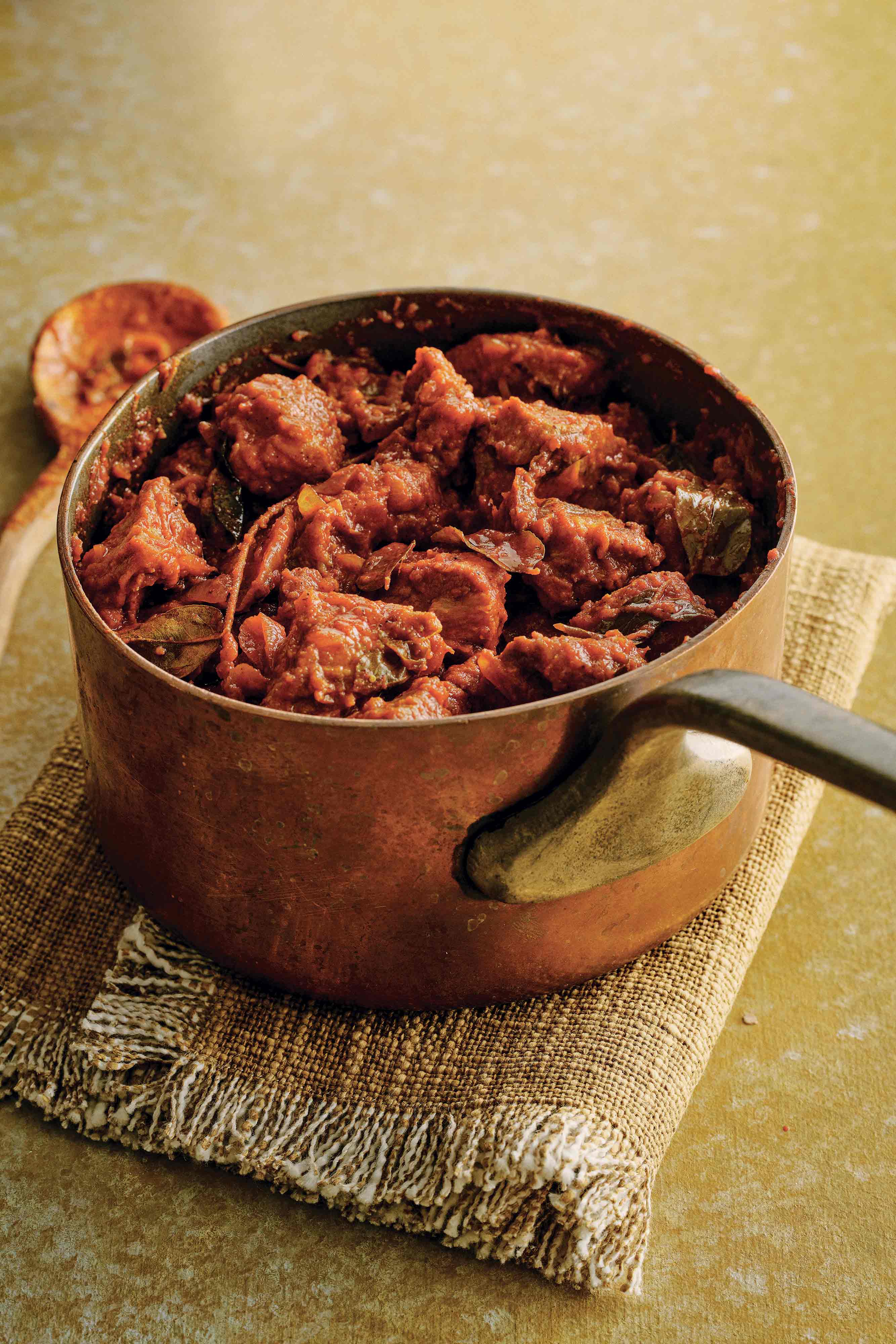 A pot with pork vindaloo in it, resting on a fringed kitchen towel with a serving spoon beside it.