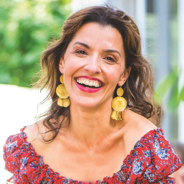 A portrait of a smiling Iranian woman outside in a red dress leaning over several dishes, with a table of people behind her.