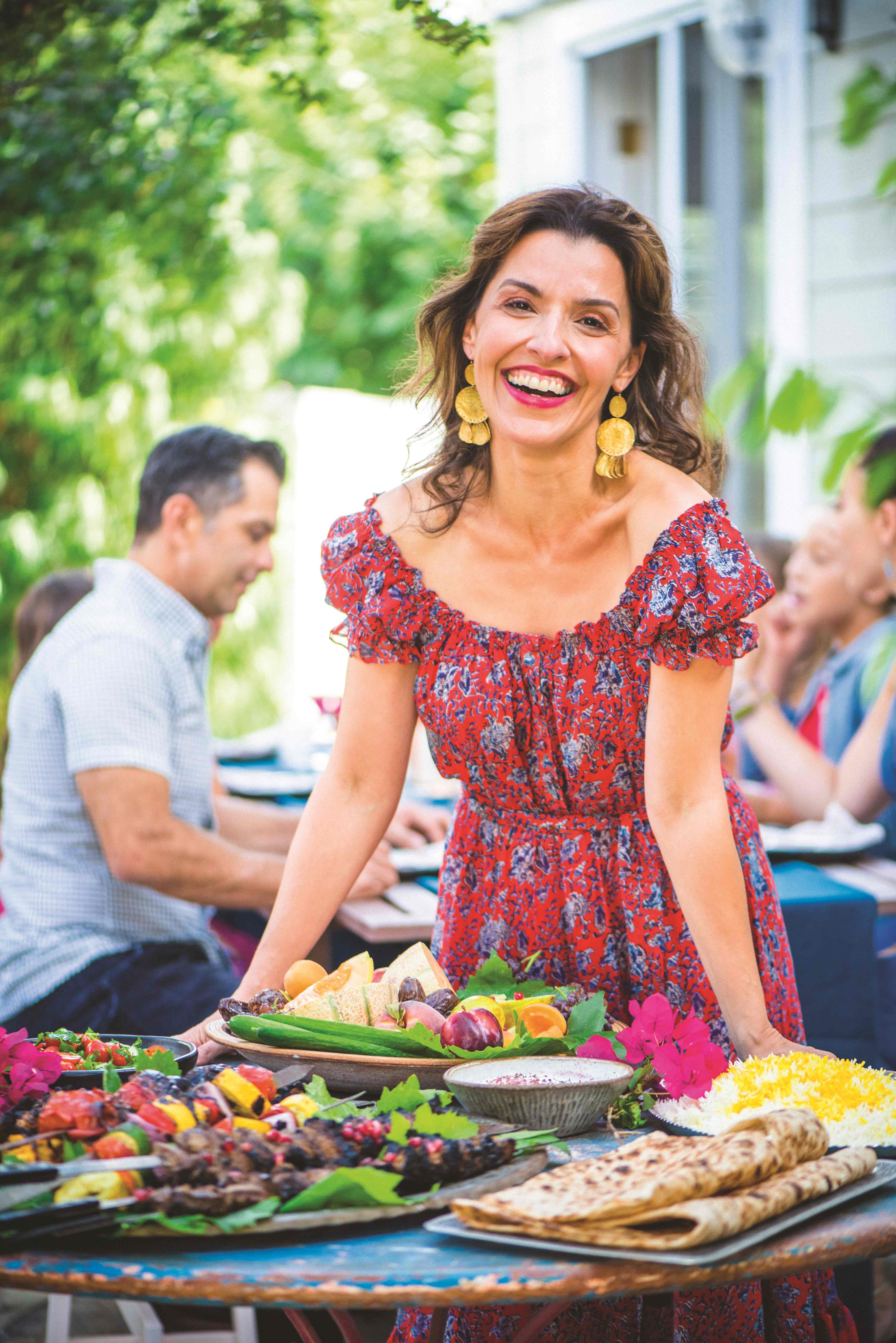 A portrait of a smiling Iranian woman outside in a red dress leaning over several dishes, with a table of people behind her.