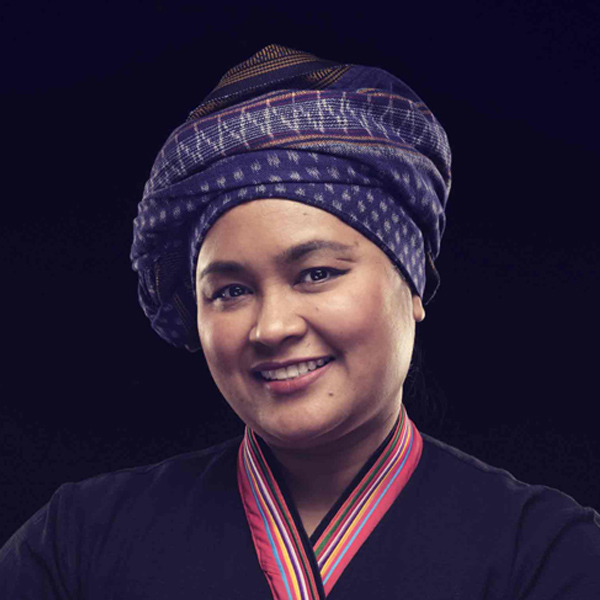 A portrait of a smiling Thai woman wearing a traditional headwrap and holding a grey mortar and pestle.