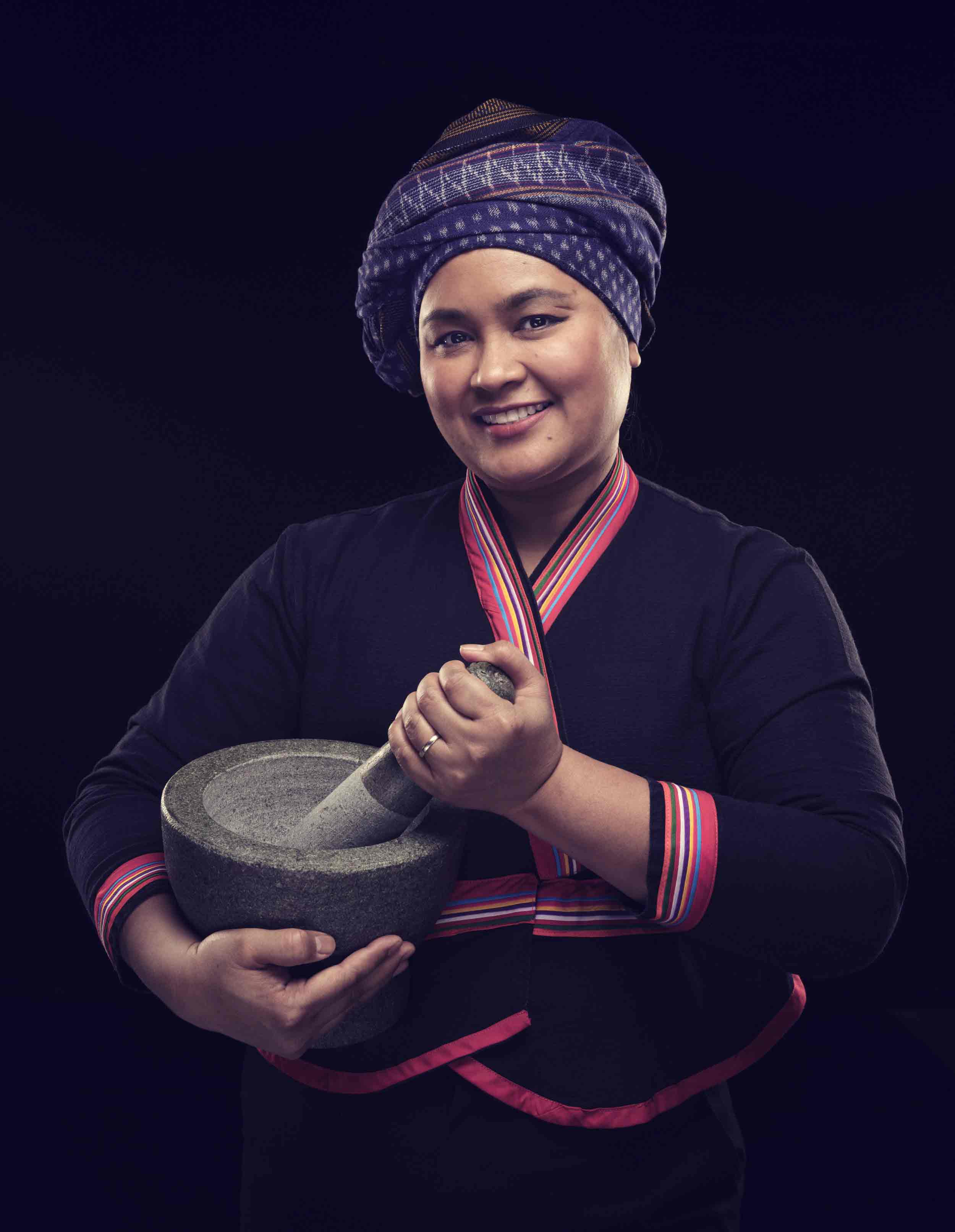 A portrait of a smiling Thai woman wearing a traditional headwrap and holding a grey mortar and pestle.