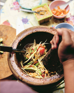 Papaya salad in a brown mortar, being mixed with a pestle and serving spoon, with ingredients in the background.