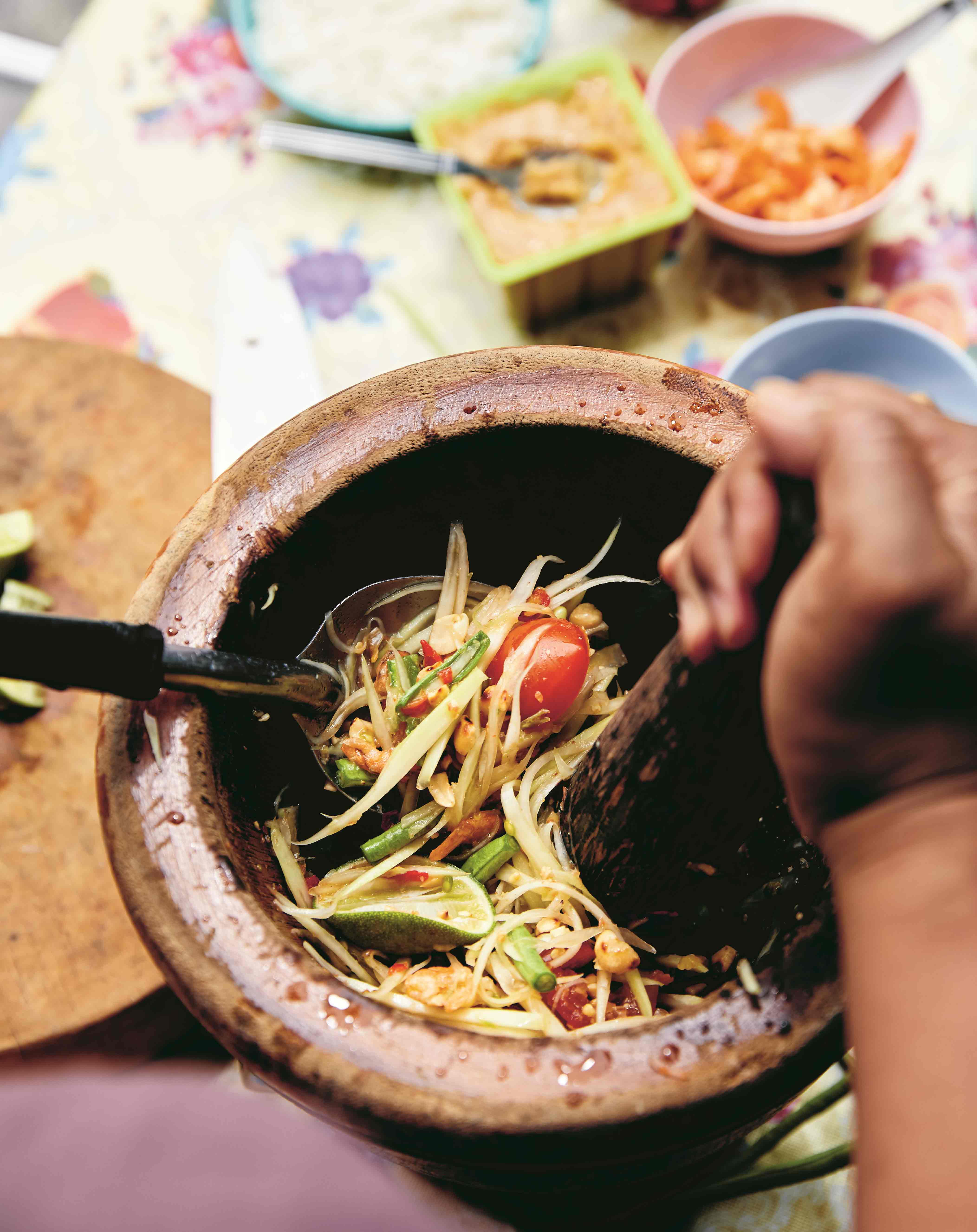 Papaya salad in a brown mortar, being mixed with a pestle and serving spoon, with ingredients in the background.