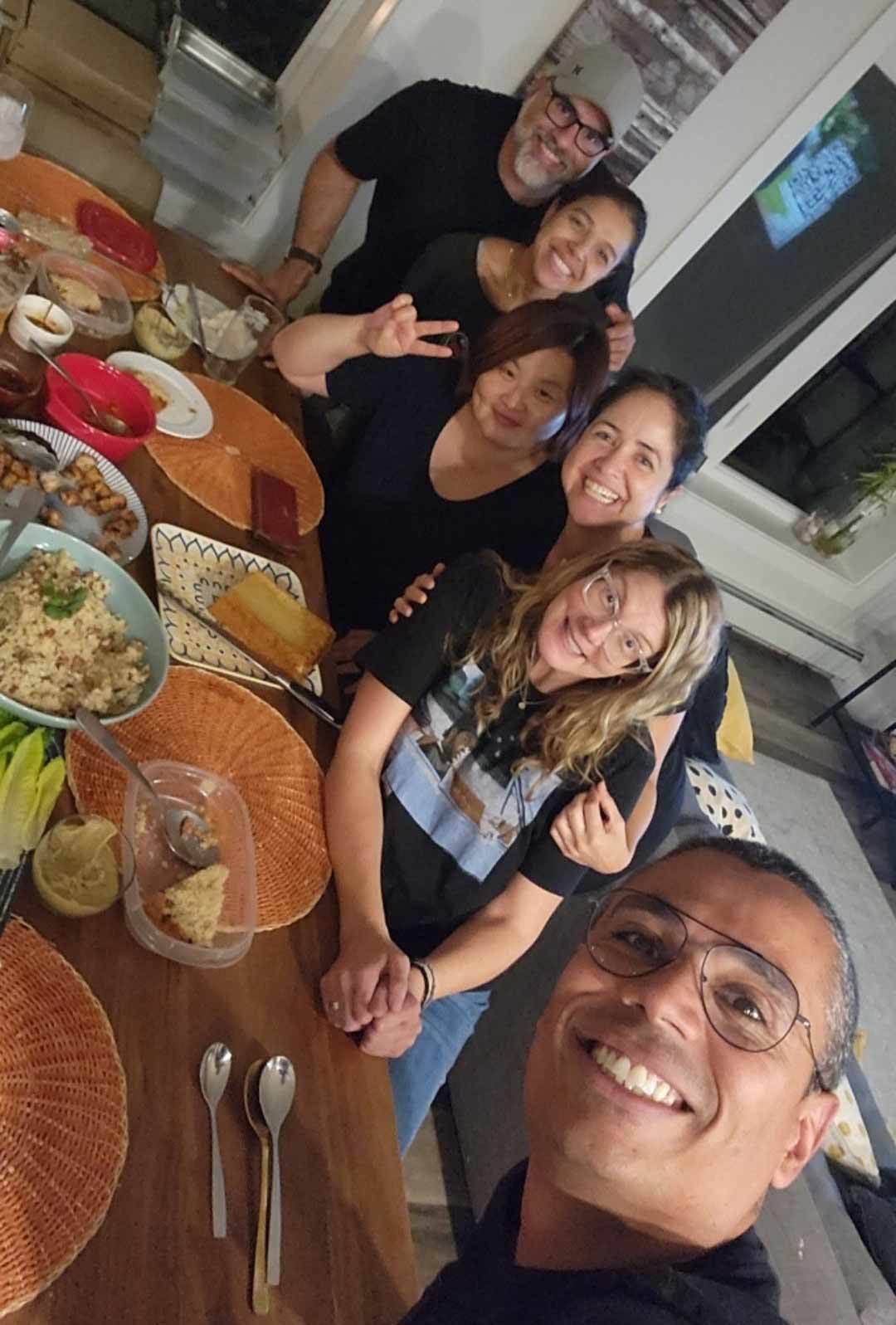A group selfie taken by a smiling man in glasses, accompanied by five other people along a wooden dining table, with food on some plates and empty plates visible from a finished meal.