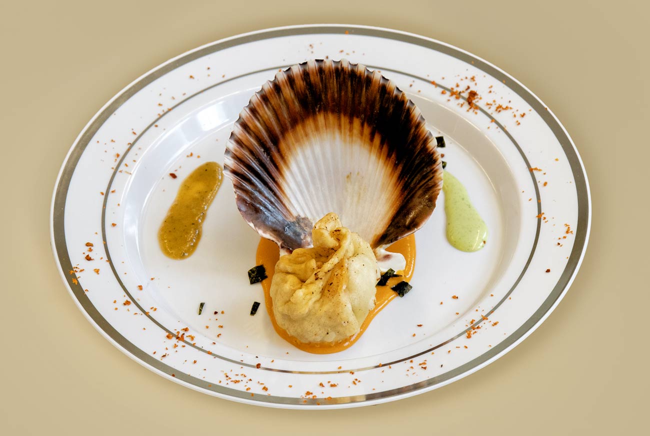 A seafood momo placed in front of a scallop shell on a white plate, set on an orange sauce base, with garlic cilantro sauce on the left and timur chutney sauce on the right.