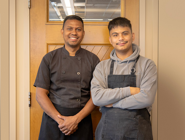 Two chefs, Darwin and Rohit, stand smiling in front of a wooden wall and door.