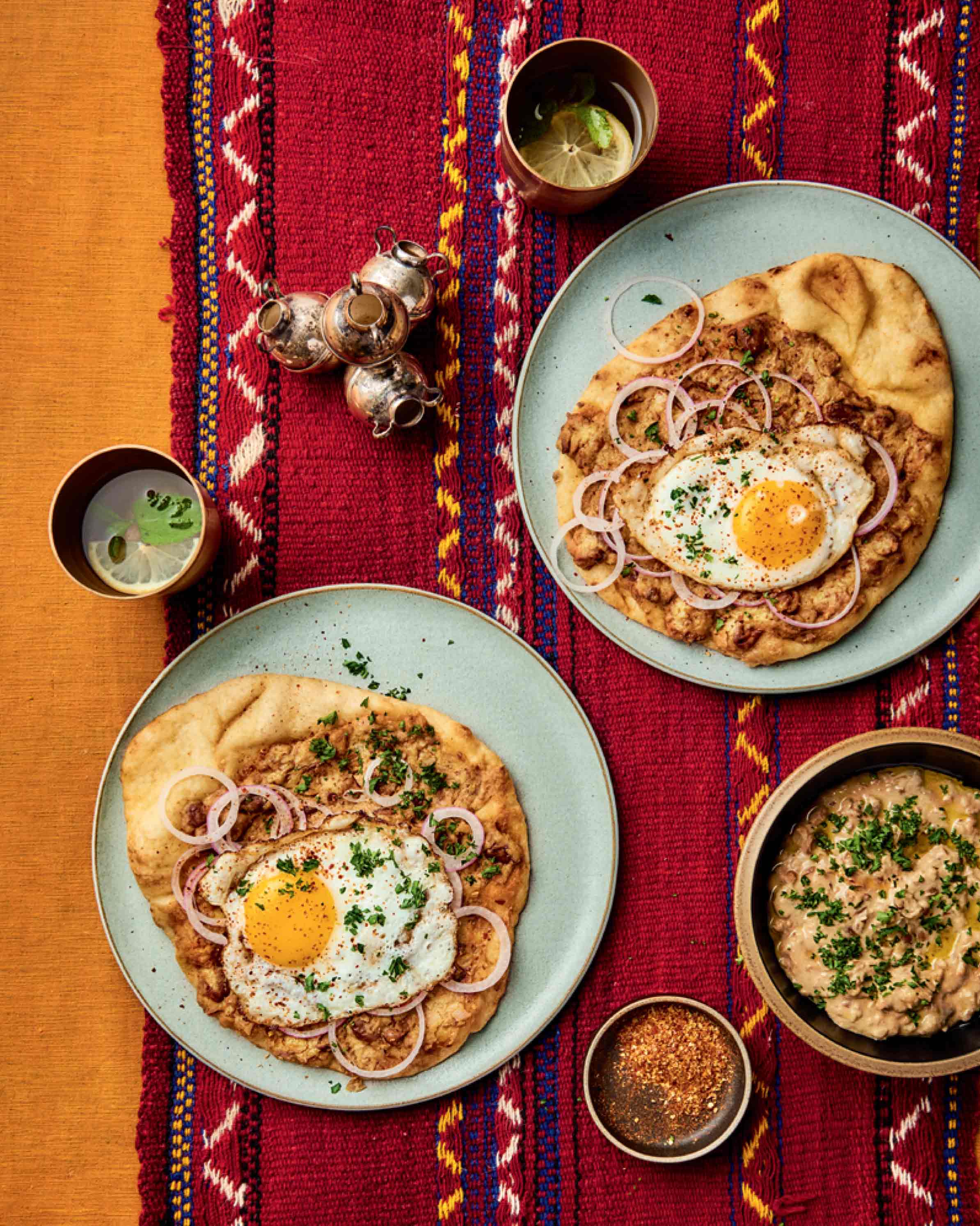 Two plates of pita, egg, and onion, ful mudammas in a bowl, and two drinks in cups, all sitting on a red decorated cloth.