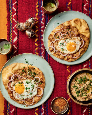 Two plates of pita, egg, and onion, ful mudammas in a bowl, and two drinks in cups, all sitting on a red decorated cloth.