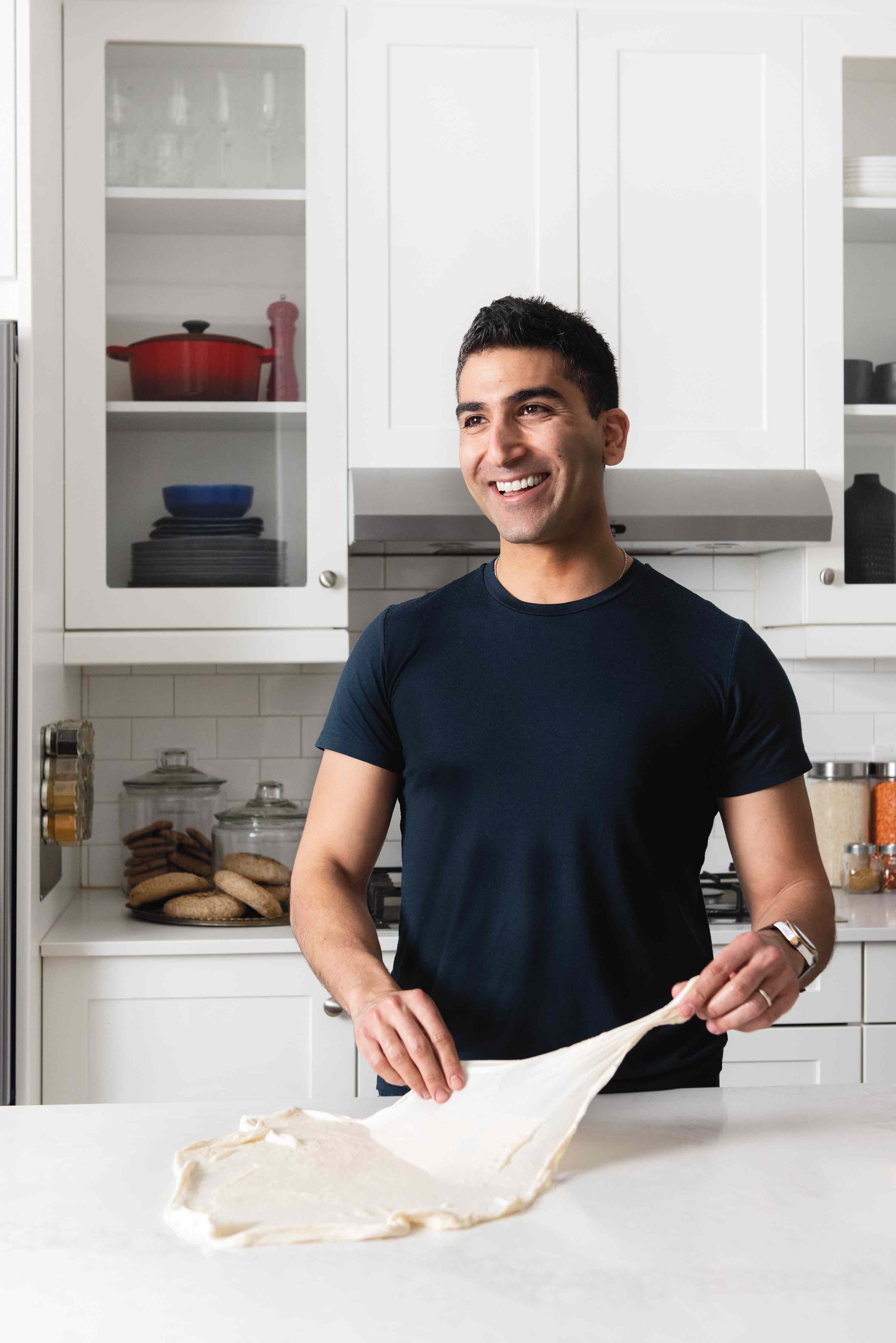 A smiling young man of Egyptian descent folding dough in a bright, white kitchen.