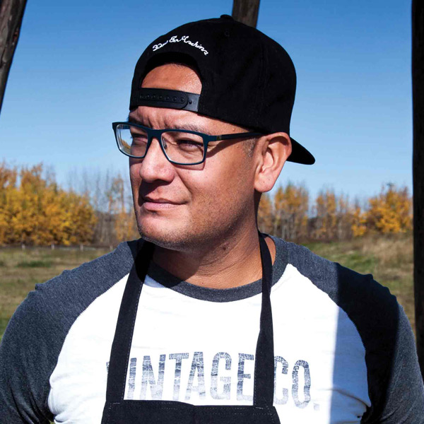 An Indigenous man wearing an apron, tshirt, and backwards ball cap, standing outside with grass and trees behind him.