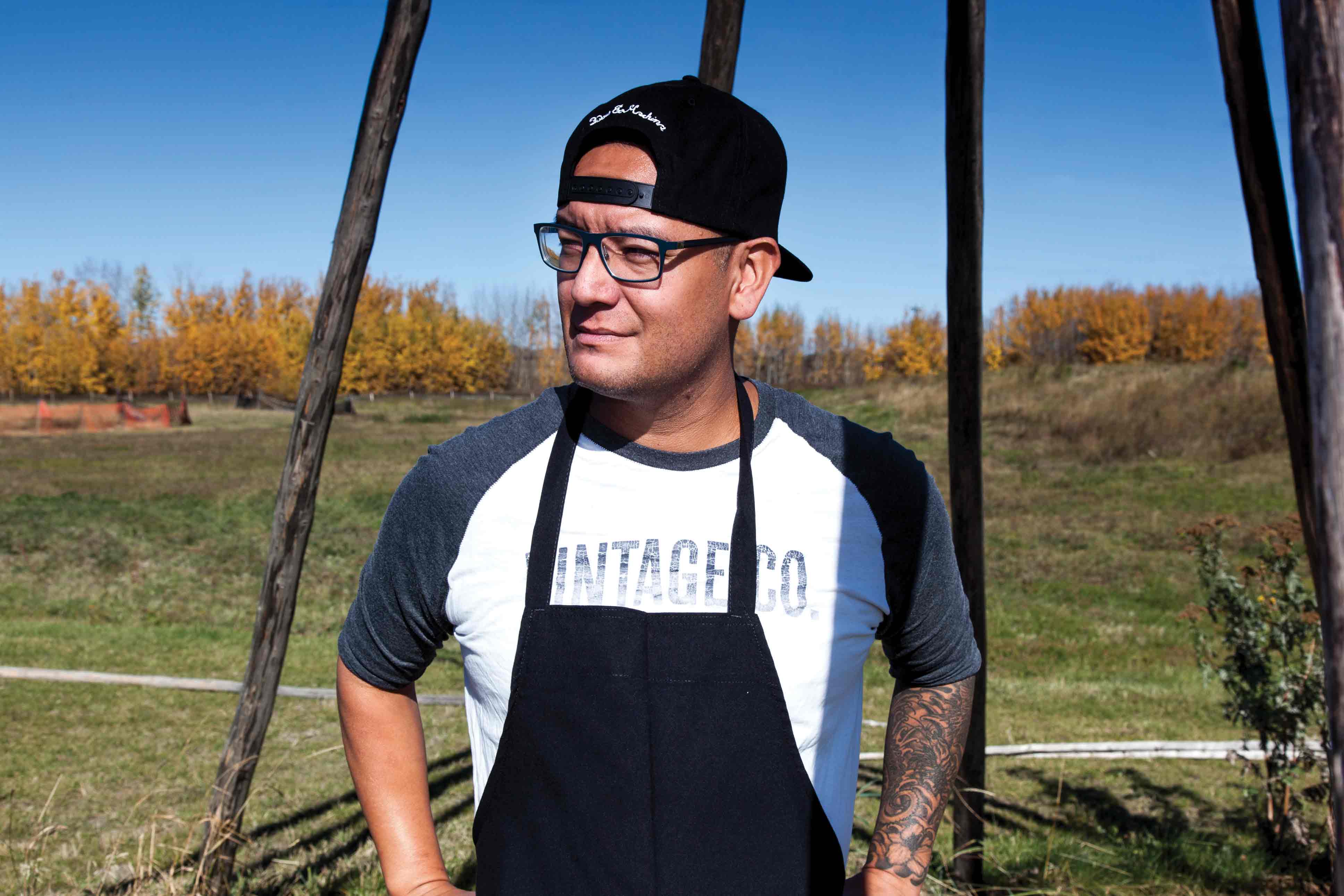 An Indigenous man wearing an apron, tshirt, and backwards ball cap, standing outside with grass and trees behind him.