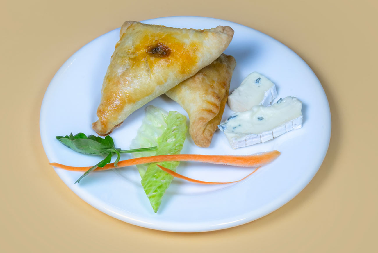 A white plate holding two golden-brown Ukajas alongside thin carrot sticks, a lettuce leaf, a sprig of parsley and two wedges of white cheese.