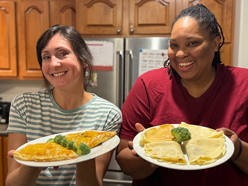 Chefs Celia and Karyna smile warmly in a kitchen while presenting freshly prepared Ukajas on large white plates.