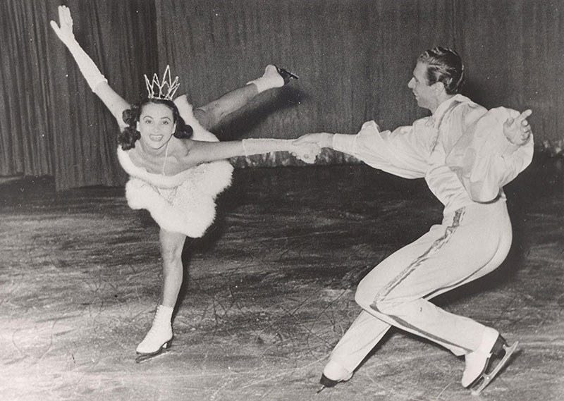 A black and white photo of a man figure skating with a woman who is wearing a small crown with her leg up in the air.