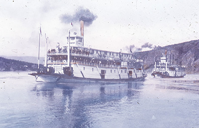 Two sternwheelers with black smoke pluming are sailing along a river.