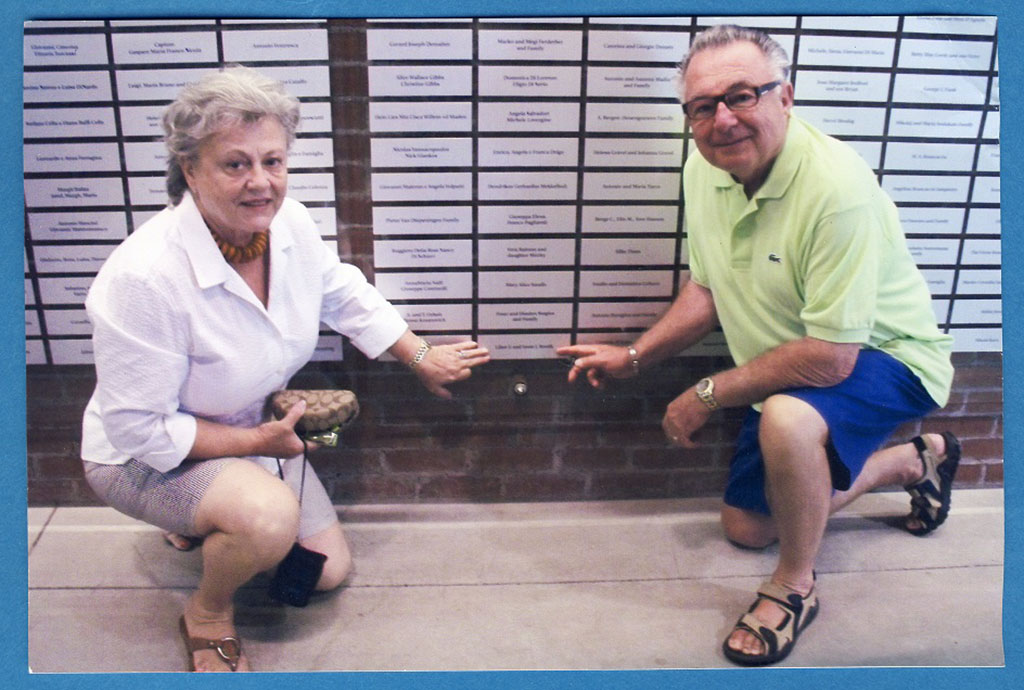A man and woman kneel before a wall of mounted plaques, they are both pointing to one of the plaques.