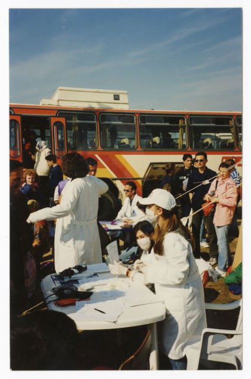 People disembark a bus, two women seated at a table are wearing white medical masks.