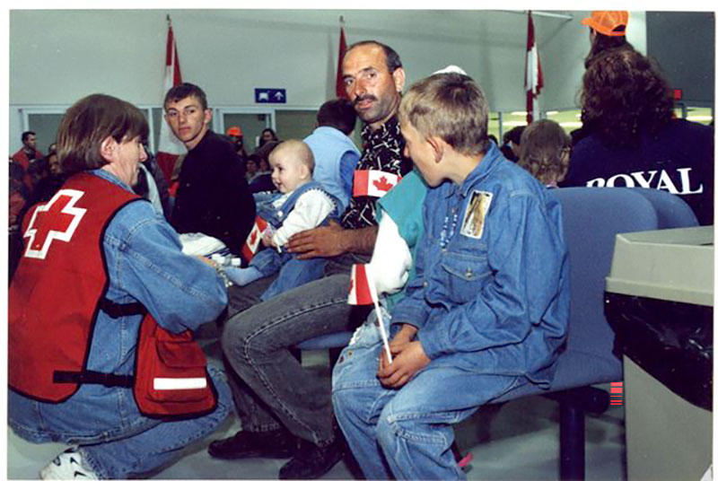 A seated man holds a baby on his lap, and the baby is holding a small Canadian flag and smiling at a woman wearing a red cross vest.
