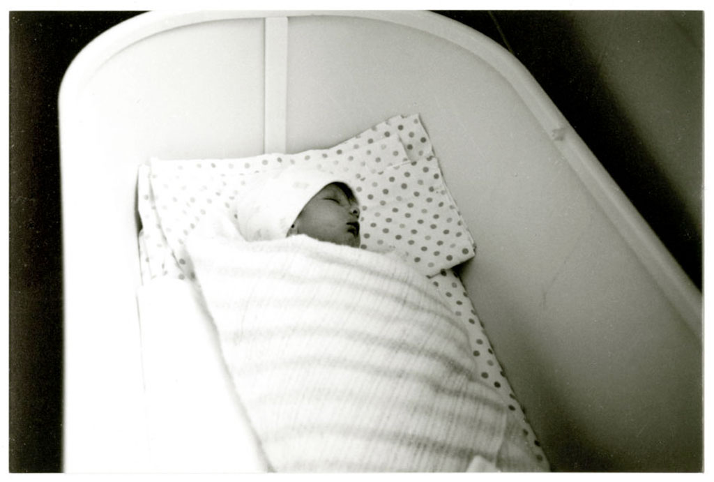A baby swaddled in a blanket sleeps in a white bassinet.