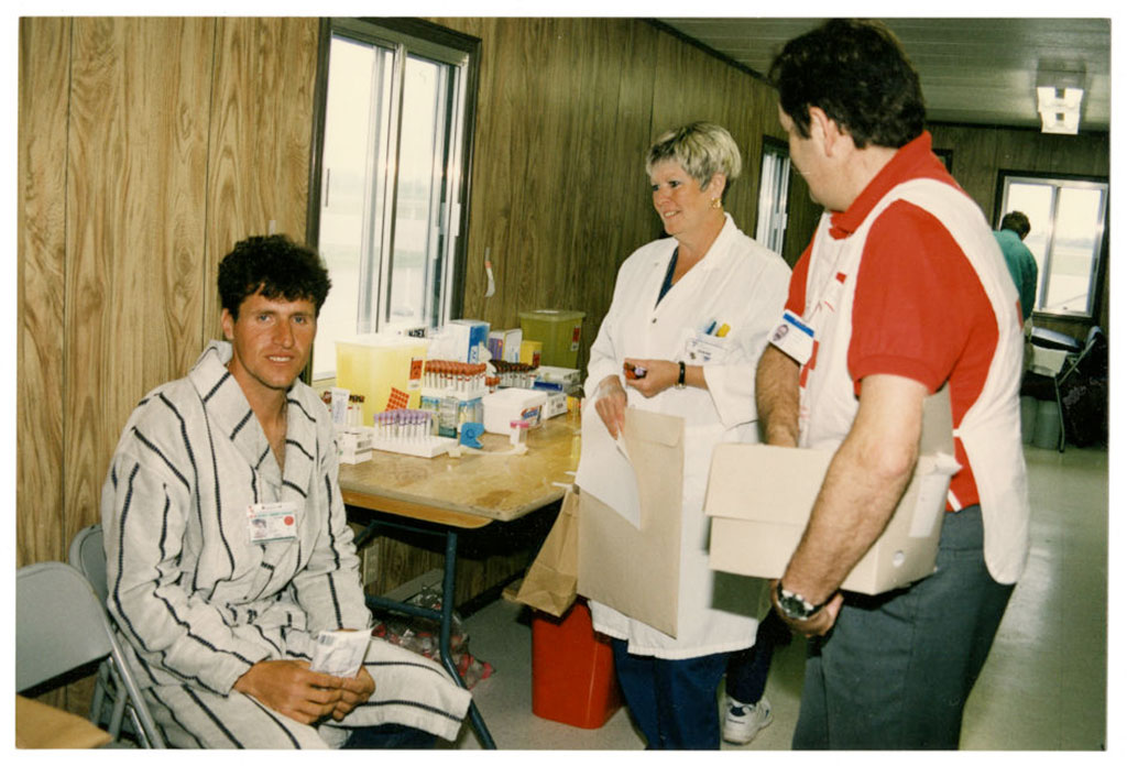 A seated man in a striped house coat smiles, while a man in a white vest and woman in a white lab coat stand next to him.