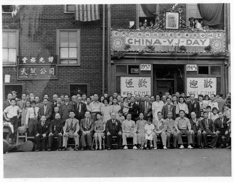 A large group of people pose outside of a building with a banner that says “CHINA-VJ-DAY.”