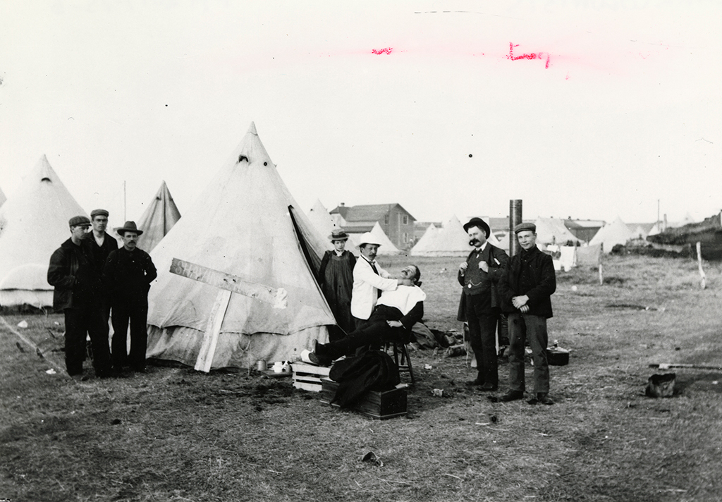 Close up of one of hundreds of tents in a work camp; men and women stand next to the tent while a barber prepares a man for shaving.