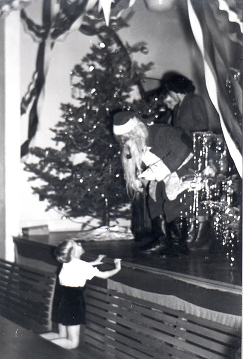 A man dressed as Santa Claus stands on a stage next to a Christmas tree and leans down to talk to a little boy on the floor.