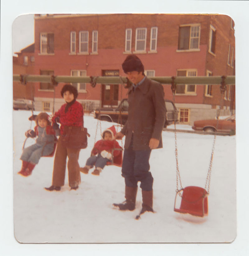 Vintage photo of a man and woman each pushing a child on a swing set. Snow covers the ground and they are in front of a large brick building.