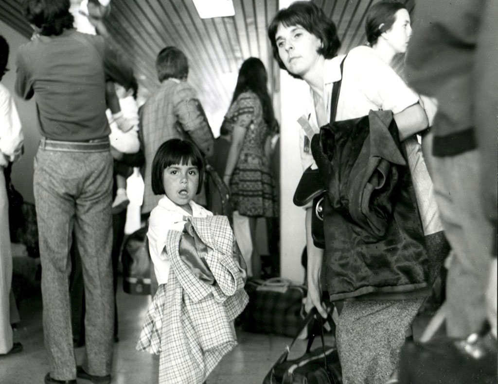 Black and white image of a dark haired child looking at the camera with her coat over her arm, next to her is a dark haired woman with a coat over her arm and holding a duffle bag. They are surrounded by other people with their backs to the camera.