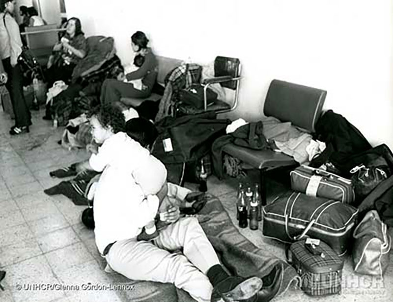 Black and white image of a room with a tile floor, a row of chairs along the wall. Luggage and clothing is scattered around and seated on a blanket the floor is a man with a baby over his shoulder.