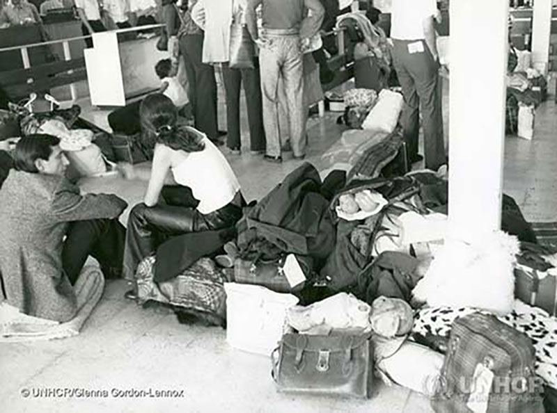 Black and white image of a room with a tile floor, a row of chairs along the wall. Luggage and clothing is scattered around and seated on a blanket the floor is a man with a baby over his shoulder.