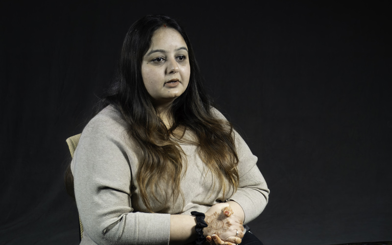 South Asian woman with long hair and beige top sitting against dark grey background.