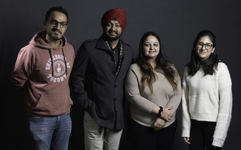Four South Asian adults, including two men, one wearing a turban, and two women standing against a dark grey background.