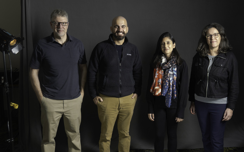 White man, Syrian man, Pakistani Woman, white-latinx woman standing against grey backdrop, beside studio light.