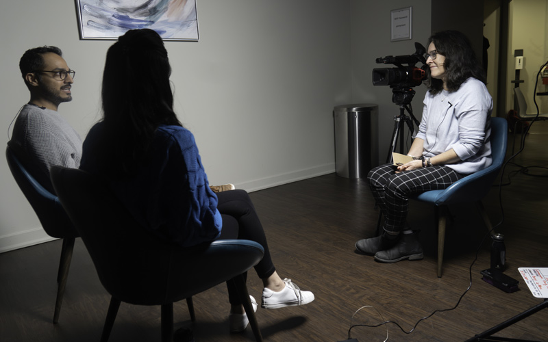 South Asian man, woman with long, dark hair seen from behind, white-latinx woman with video camera beside her, all sitting.
