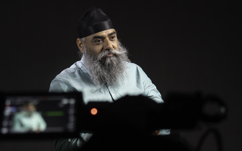 South Asian man with turban, beard and blue shirt, camera and monitor in foreground.