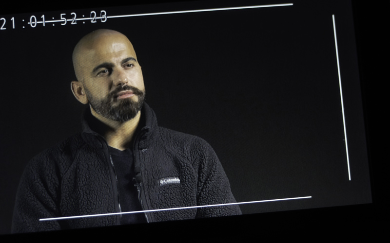 Angled video-camera monitor view of Middle Eastern man with beard, black top and dark grey jacket against black background.