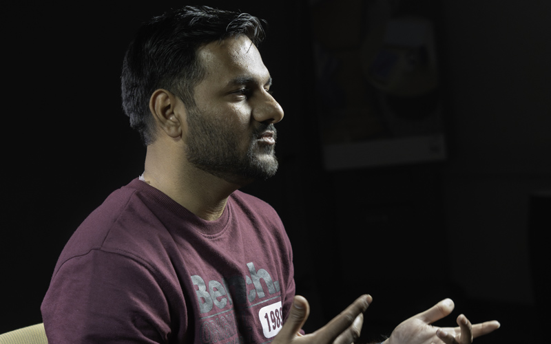 South Asian man with black hair and beard, maroon shirt, and hands in air against black background.
