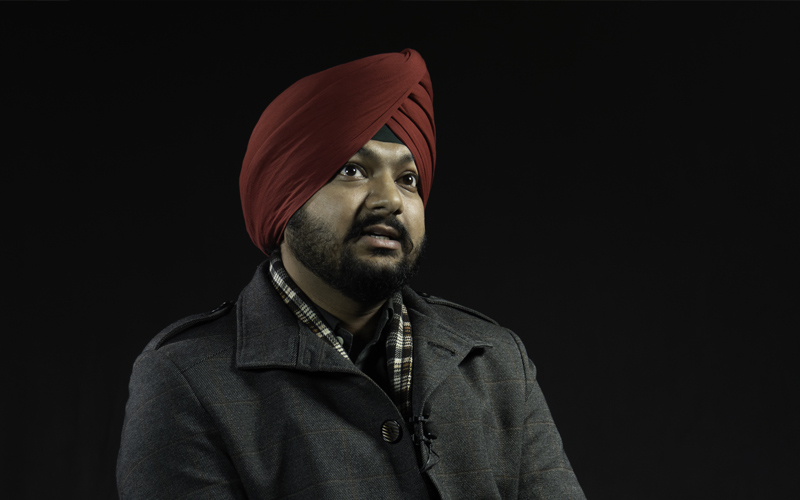 South Asian man with turban, beard, grey jacket with red lines, and scarf against black background.