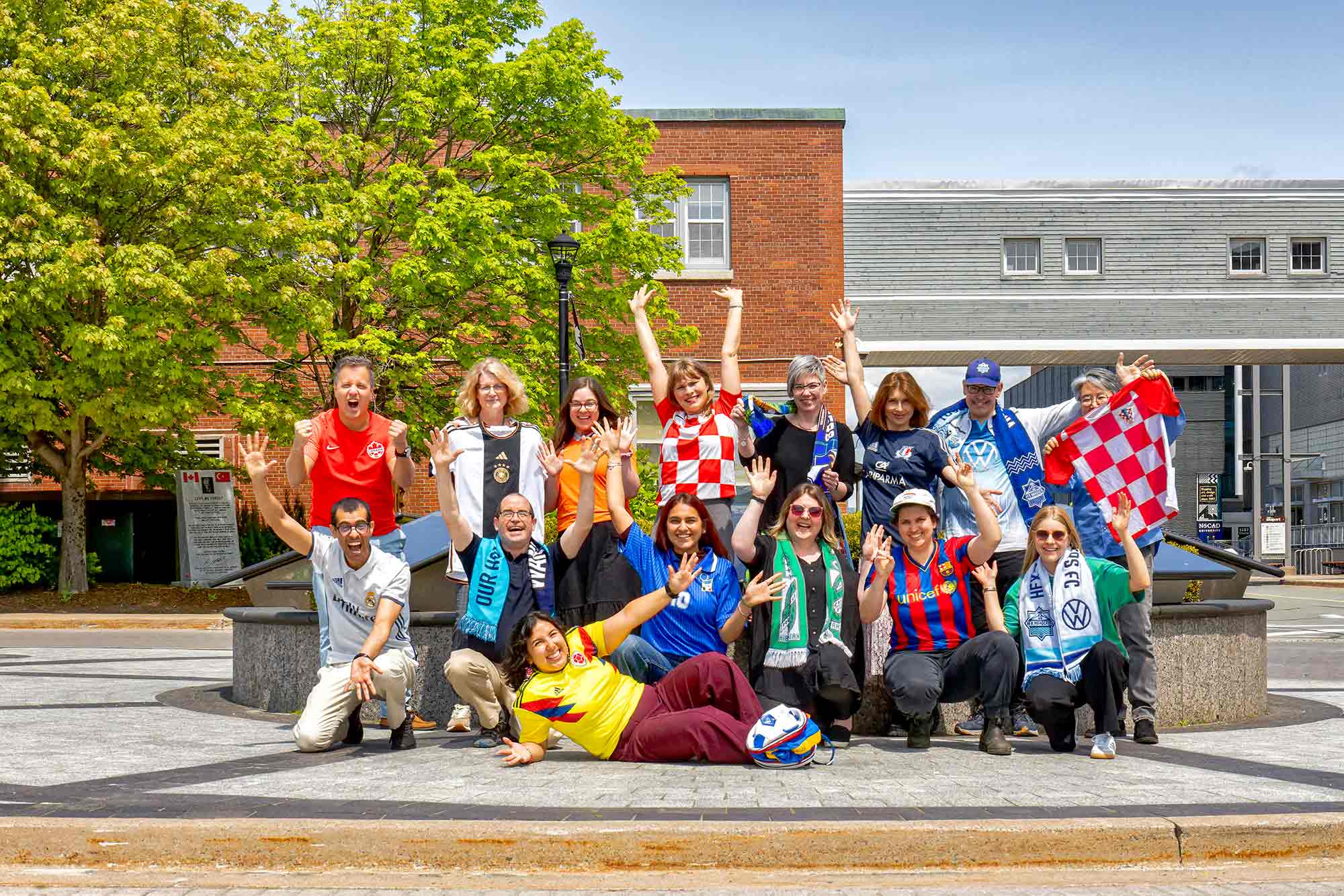 Fifteen people cheering, wearing various soccer teams, in two rows with one woman lying on the ground at front.