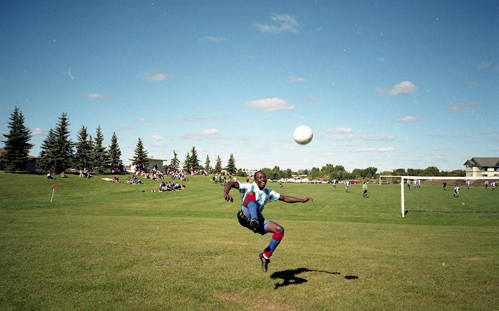 A black man smiles mid-air after kicking a ball, in bright sun on a large field with an active soccer pitch behind him.